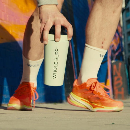 Wearing Whole Supp Performance Running Socks and bright orange running shoes, a person crouches on a city sidewalk, holding a white WHOLE SUPP® shaker bottle by a colorful graffiti wall.